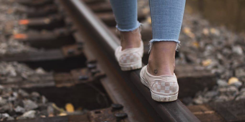 The Daughter. Photo by William Daigneault on Unsplash shows a girl in sneakers and blue jeans walking on a single train track