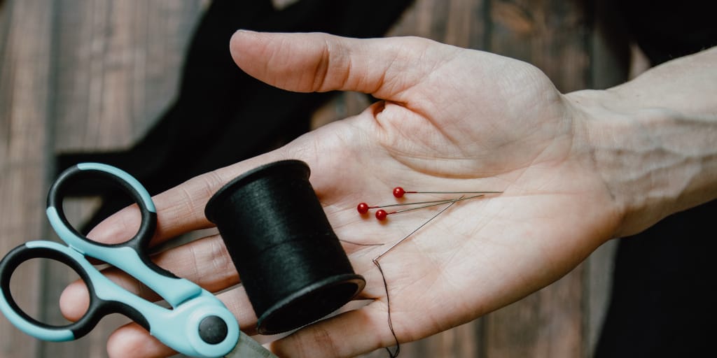 A Hole in November. Photo by Kelly Sikkema on Unsplash. Shows an open hand holding sewing pins, a reel of dark thread and scissors.