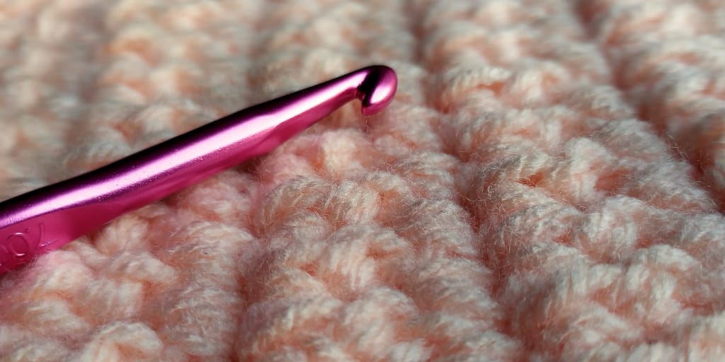 Crochetwork. Photo by Karina L on Unsplash. Shows a shining pink crochet hook on crochet worked in peach yarn.